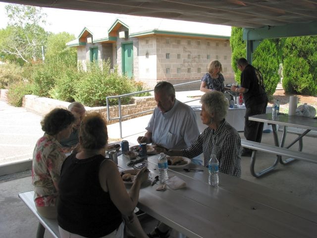 August 2015 Picnic — members dining at picnic tables under covered pavilion with stone restroom building and trees visible in background
