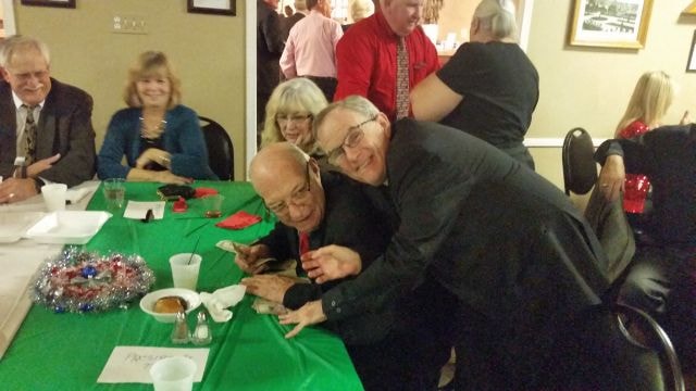 December 2015 Christmas Dinner — two men leaning together smiling at green-tablecloth table with guests behind them